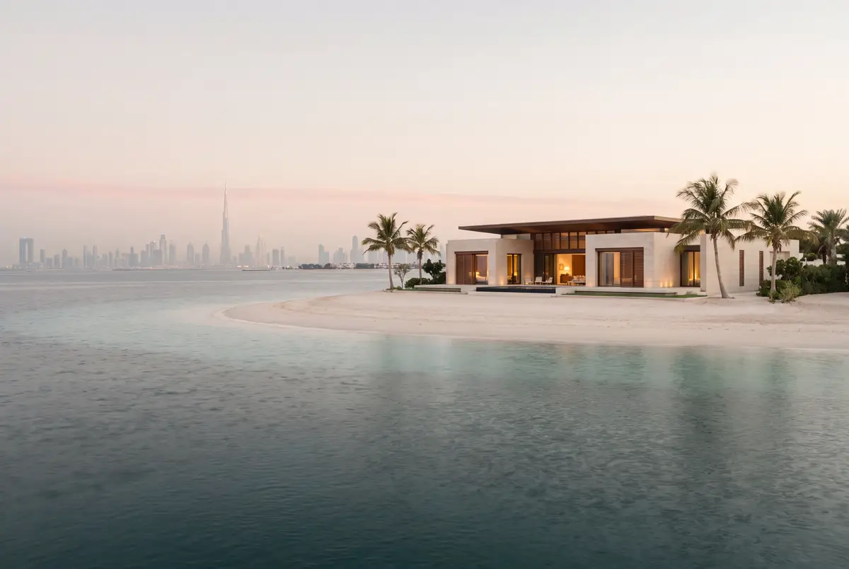A single-storey villa among palms on a small white-sand island, with the Dubai skyline faint on the horizon